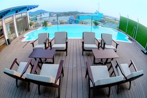 A rooftop pool area with lounge chairs and tables, overlooking a cityscape.