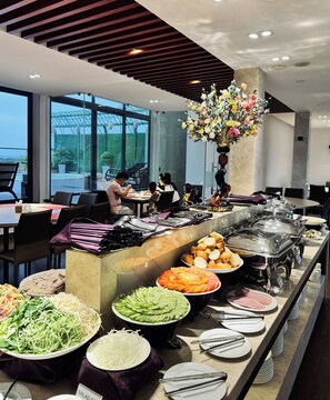 Montaje de bufé con diversos alimentos y utensilios, un jarrón grande con flores y una zona de estar con personas cenando.