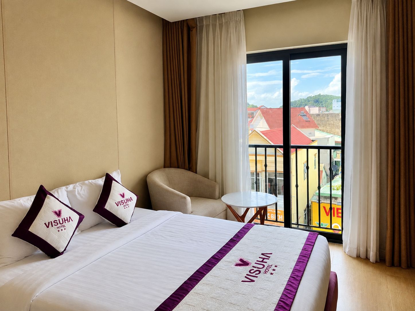 Habitación de hotel con una cama, una silla, una mesita y un balcón con vistas a edificios.