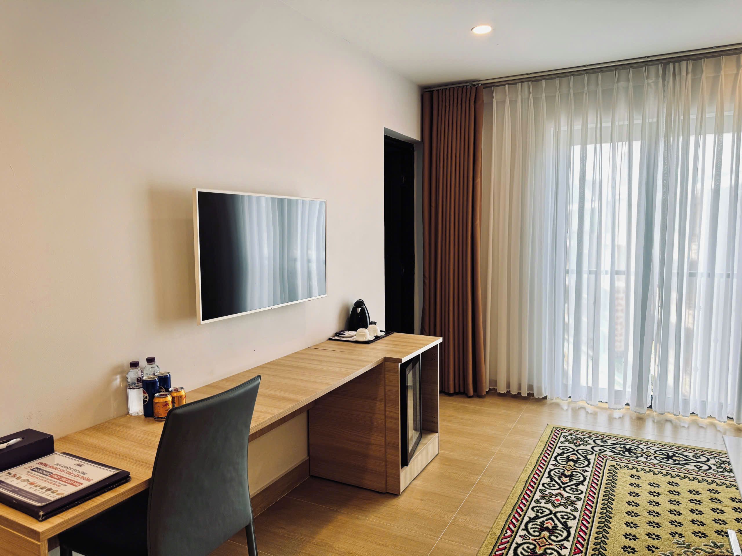 A modern hotel room with a flat-screen TV mounted on the wall, a wooden desk with a chair, a coffee maker, and a patterned rug on the floor.