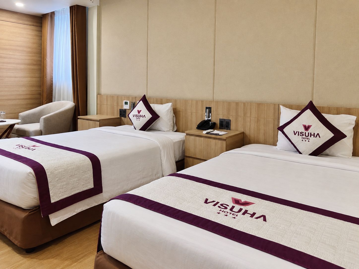 Habitación de hotel con dos camas, cada una con almohada con el logotipo de la marca y cabecero. Se aprecia una mesita de noche con teléfono y lámpara.