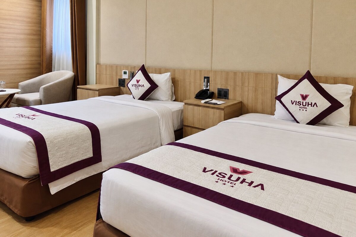 A hotel room with two beds, each with a branded pillow and a headboard. There is a bedside table with a phone and a lamp.