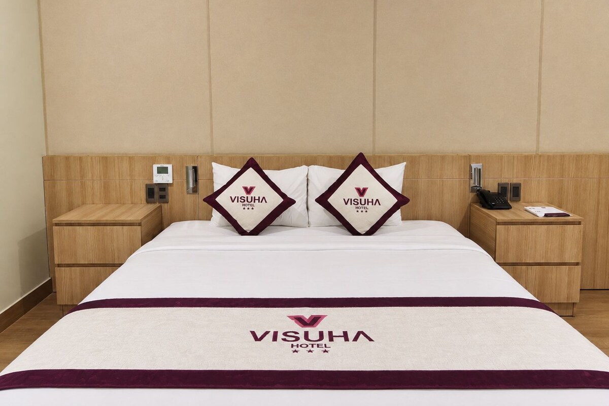 A hotel room with a bed, two pillows with the hotel logo, a bedside table with a phone, and a control panel on the wall.