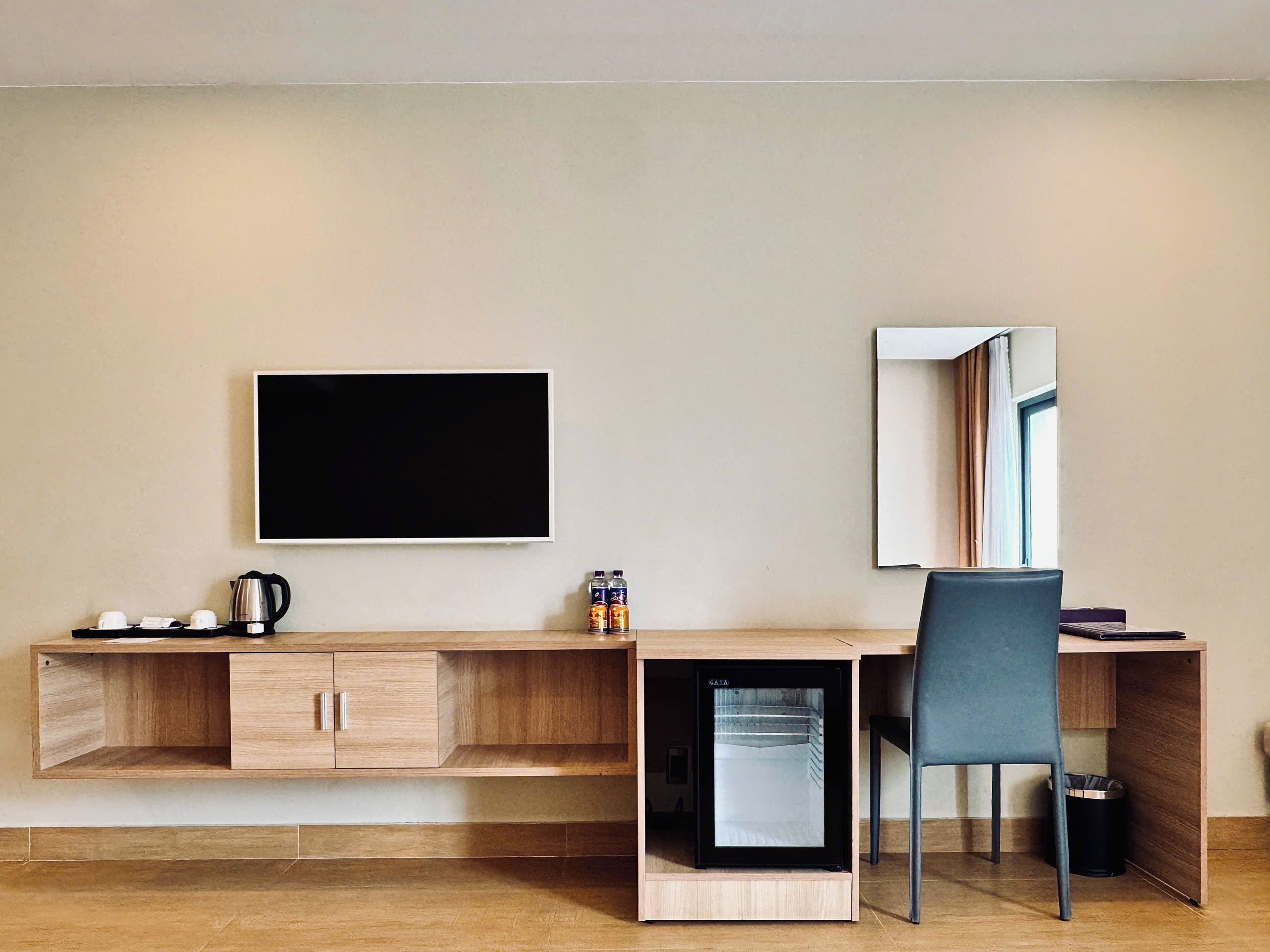 A compact hotel room with a wooden desk, a small refrigerator, a chair, a flat-screen TV, and a mirror.