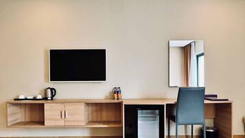 A compact hotel room with a wooden desk, a small refrigerator, a chair, a flat-screen TV, and a mirror.