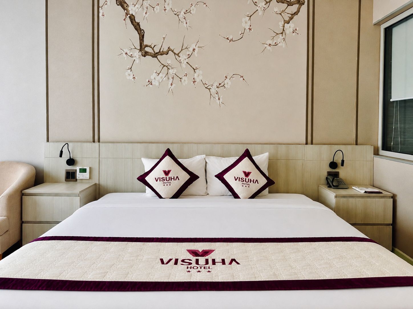 A hotel room with a bed, two pillows with the hotel logo, a nightstand with a phone, and a wall art piece.
