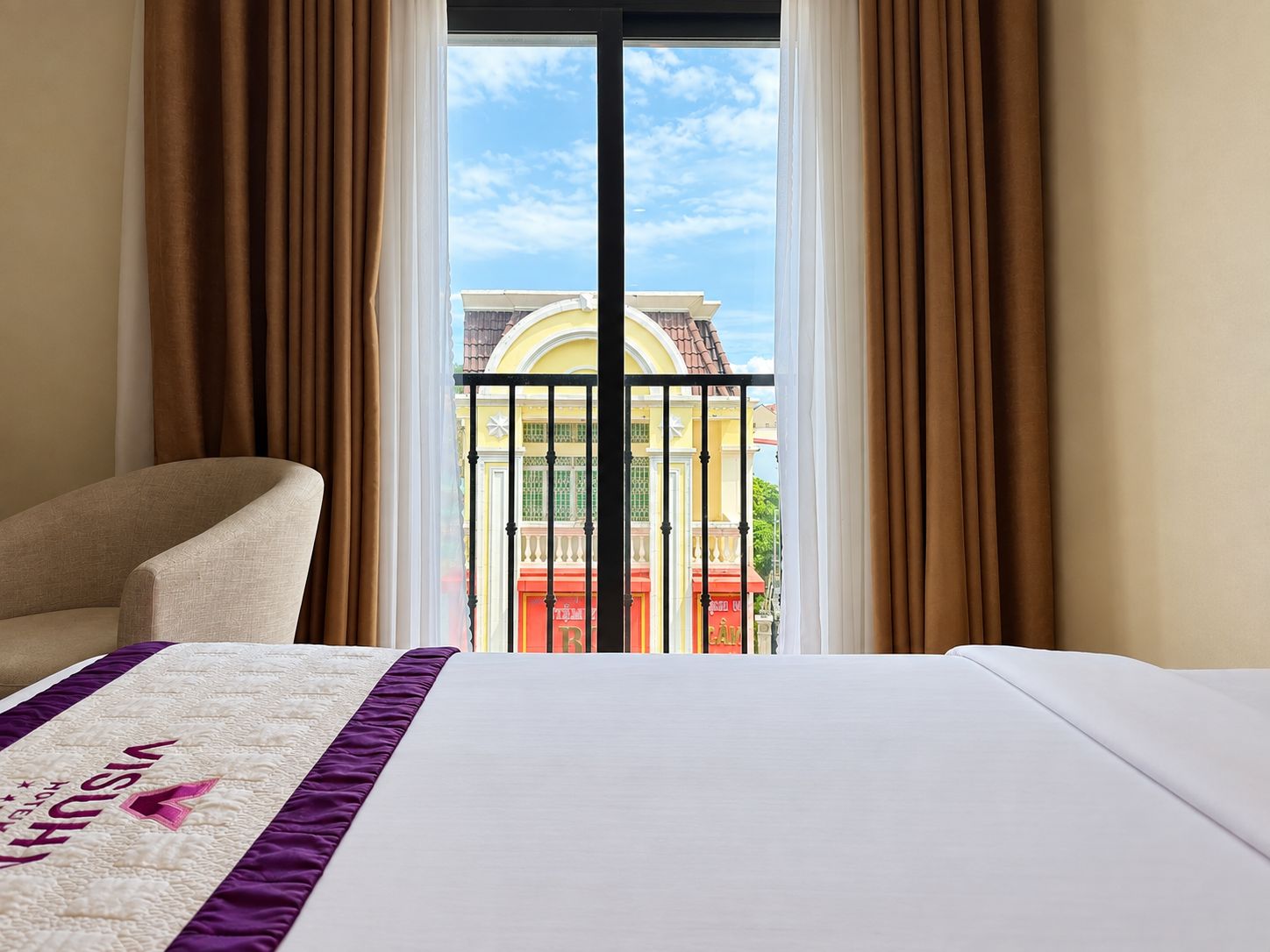 A hotel room with a bed, a chair, and a view of a building through a window with curtains.