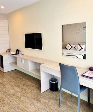 A modern hotel room with a flat-screen TV, a bed with decorative pillows, a desk with a chair, and a trash can.