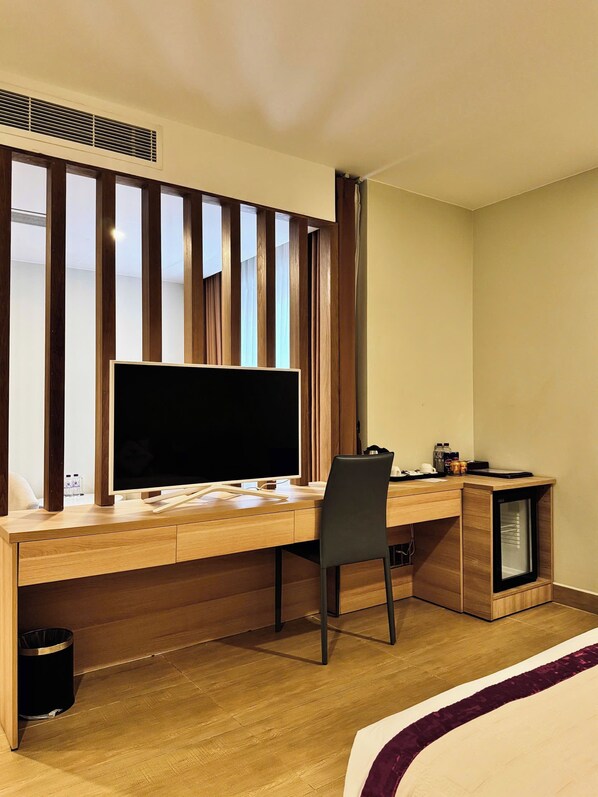 Habitación de hotel con escritorio de madera, televisor de pantalla plana grande, una silla, un refrigerador pequeño y un bote de basura.