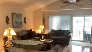 Ein Wohnzimmer mit zwei Sofas, einem Couchtisch und zwei Lampen. Es gibt ein Fenster mit Jalousien und einen Blick auf Gebäude im Freien.