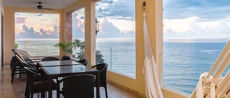 A room with a dining table and chairs, a hammock, and a view of the sea.