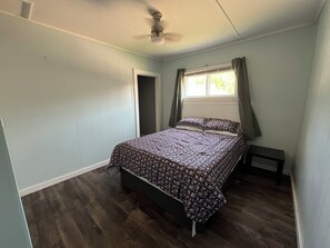 Ein Schlafzimmer mit Bett, Nachttisch, Fenster mit Vorhängen, Tür und Deckenventilator.