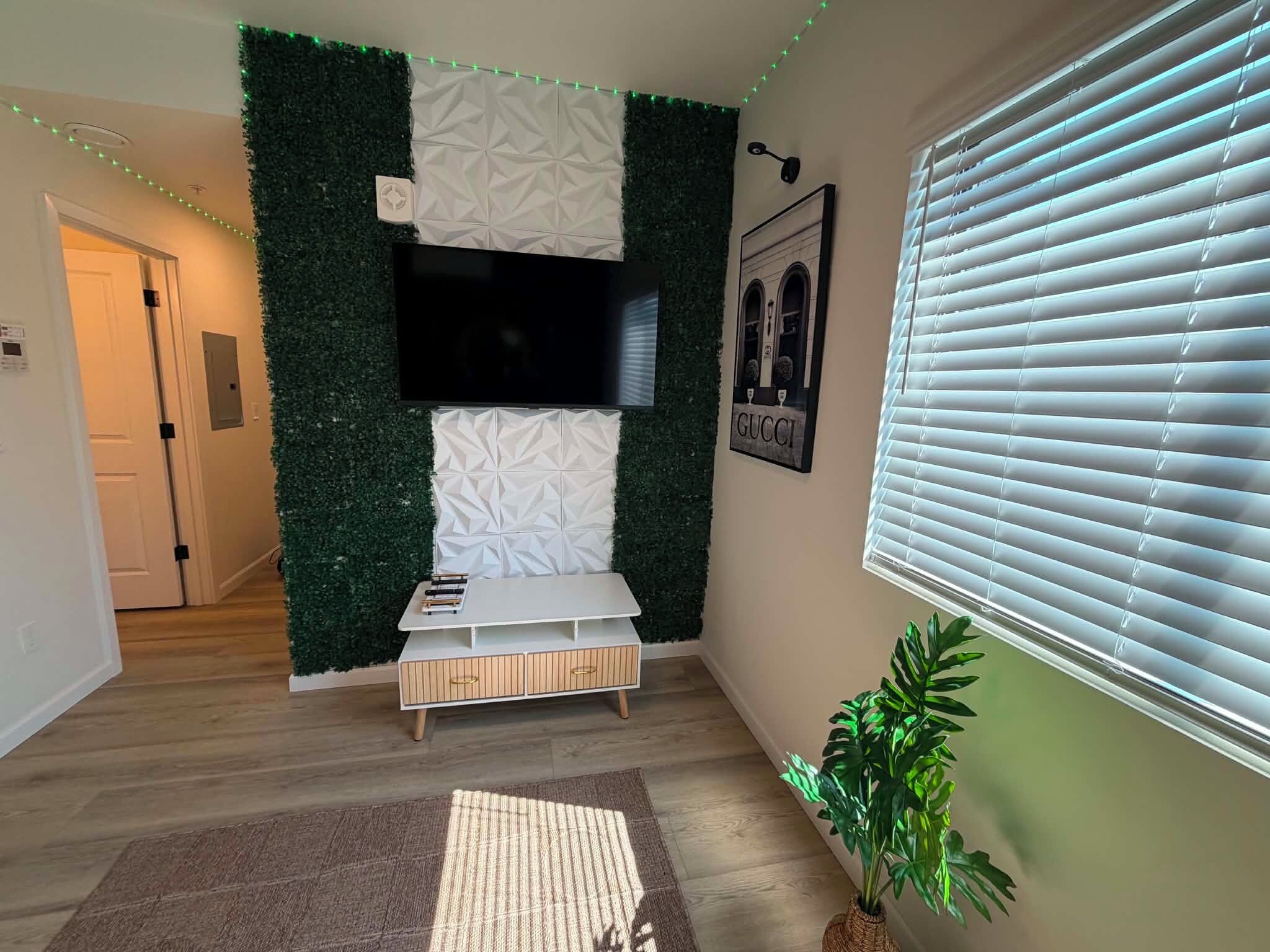 A modern living room with a flat-screen TV mounted on a textured wall, a white console table, a potted plant, and a window with blinds.