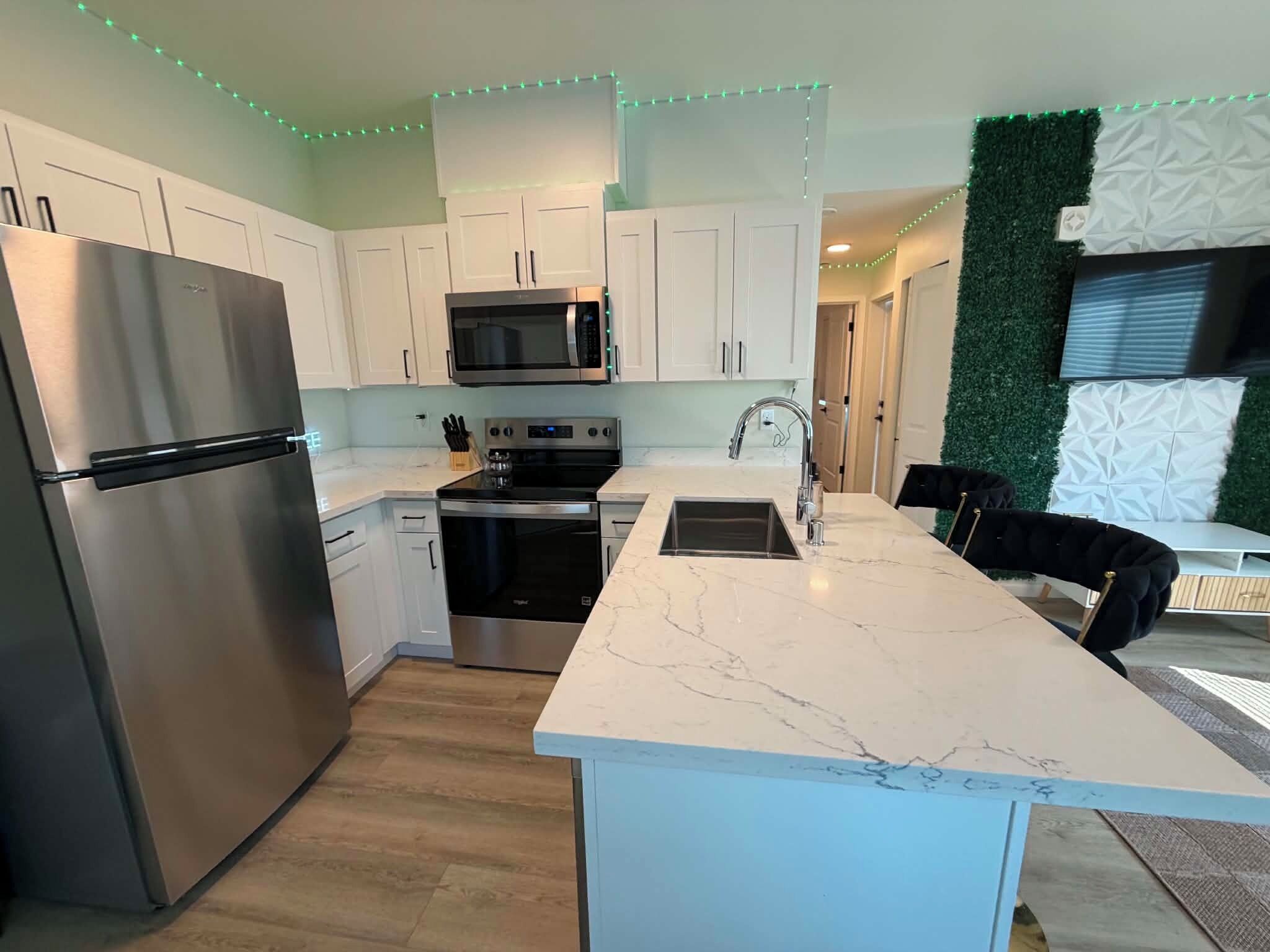 Uma cozinha moderna com armários brancos, eletrodomésticos de aço inoxidável, bancada de mármore e iluminação em LED verde.