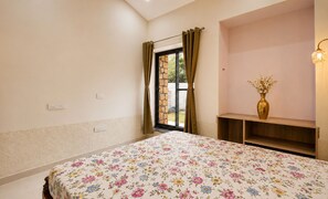 A bedroom with a floral bedspread, a window with curtains, a bedside table with a vase, and a wall-mounted shelf.