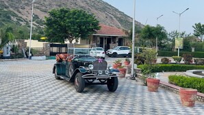 A paved area with a classic car, a building with a red roof, and a hill in the background.