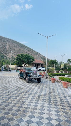A paved area with a classic car, a building with a red roof, and a hill in the background.