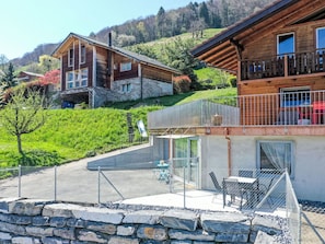 A chalet-style building with a stone foundation, a balcony with a metal railing, and a clear view of the surrounding greenery and hills.