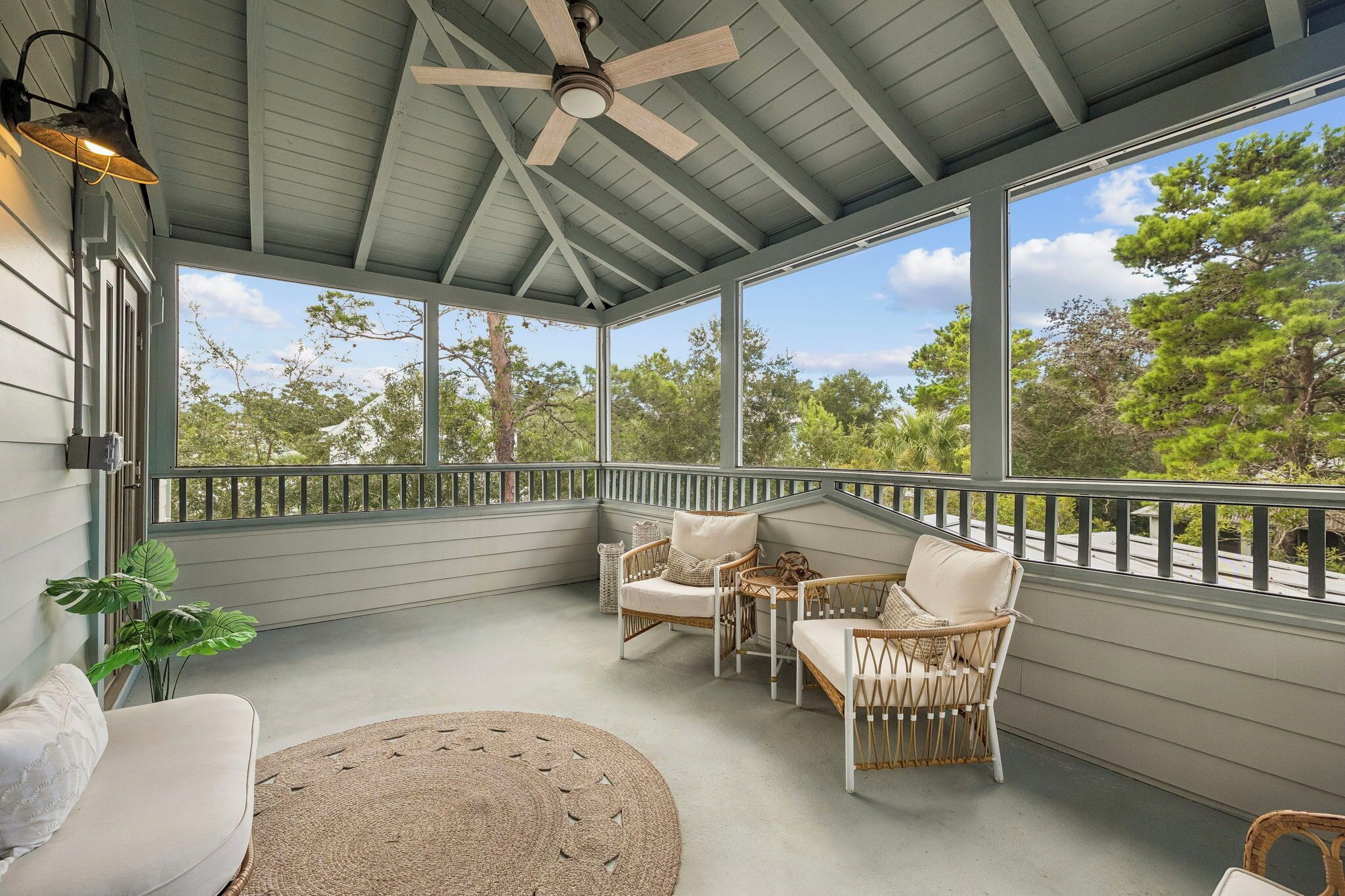 A covered porch with wicker furniture, a ceiling fan, and large windows offering a view of trees.