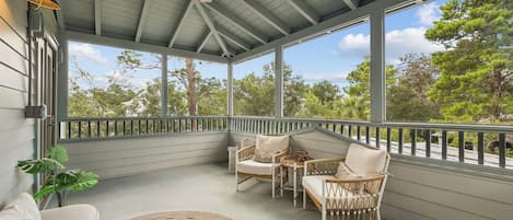 A covered porch with wicker furniture, a ceiling fan, and large windows offering a view of trees.