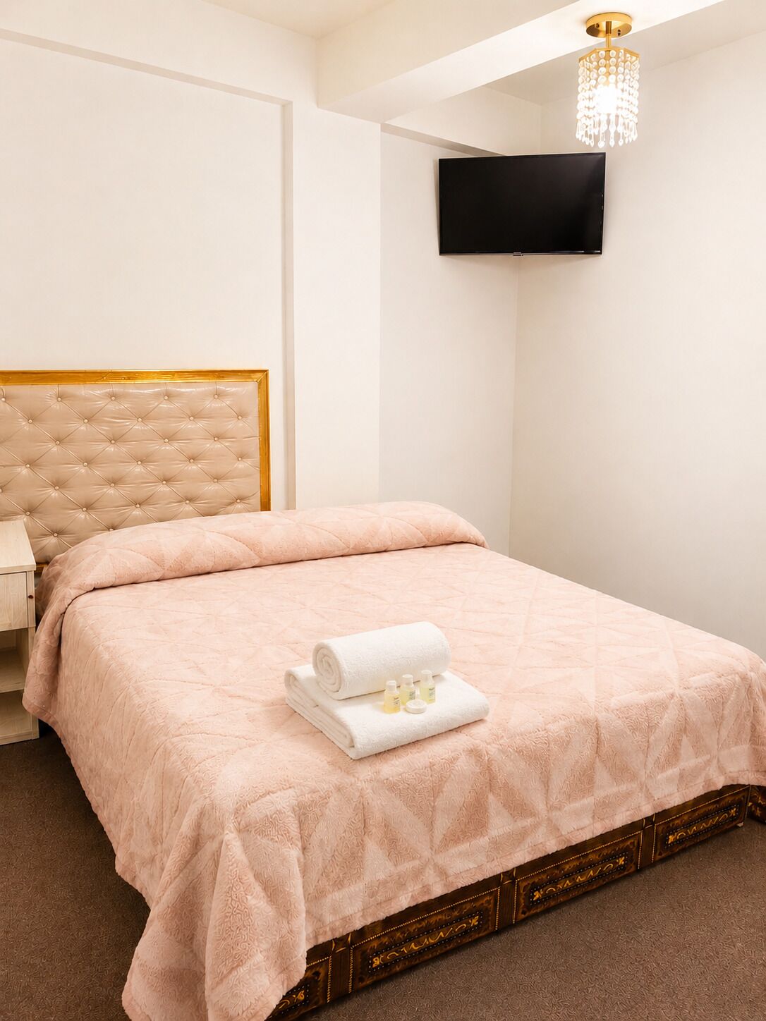 Een netjes opgemaakt bed met een opgerolde witte handdoek en toiletartikelen, een gestoffeerd hoofdbord, een flatscreen televisie aan de wand en een kroonluchter.