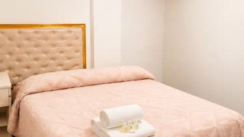 A neatly made bed with a rolled white towel and toiletries, a tufted headboard, a flat-screen TV mounted on the wall, and a chandelier.