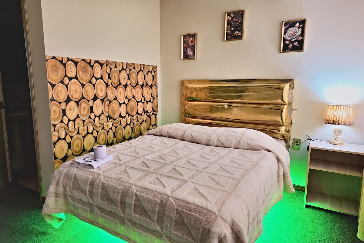 Habitación de hotel con cama, cabecera con diseño de veta de madera, mesita de noche con lámpara y suelo iluminado de color verde.