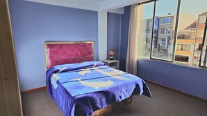 A hotel room with a bed, a nightstand, a lamp, and a window with a city view.