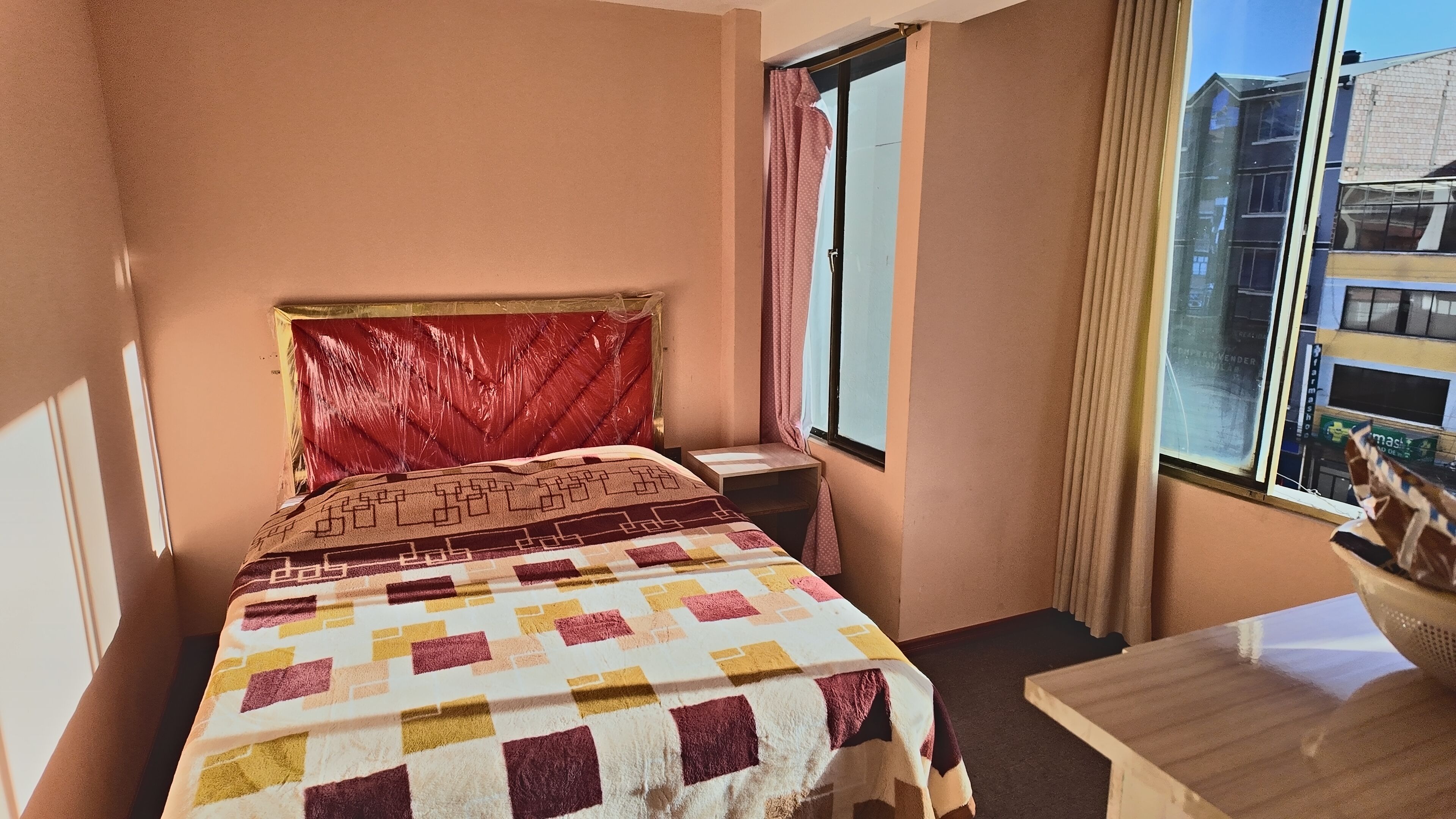 A hotel room with a bed, a small table, a window with curtains, and a view of buildings outside.