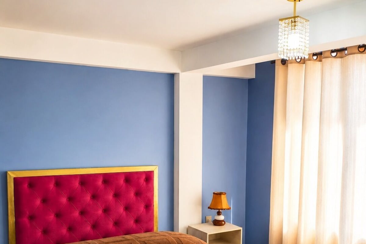 Habitación de hotel con una cama grande, cabecera, mesita de noche con lámpara y ventana con cortinas.