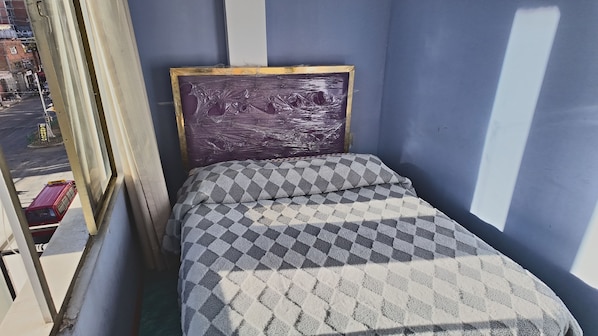 Una habitación pequeña con una cama, un ventanal y vista a la calle.