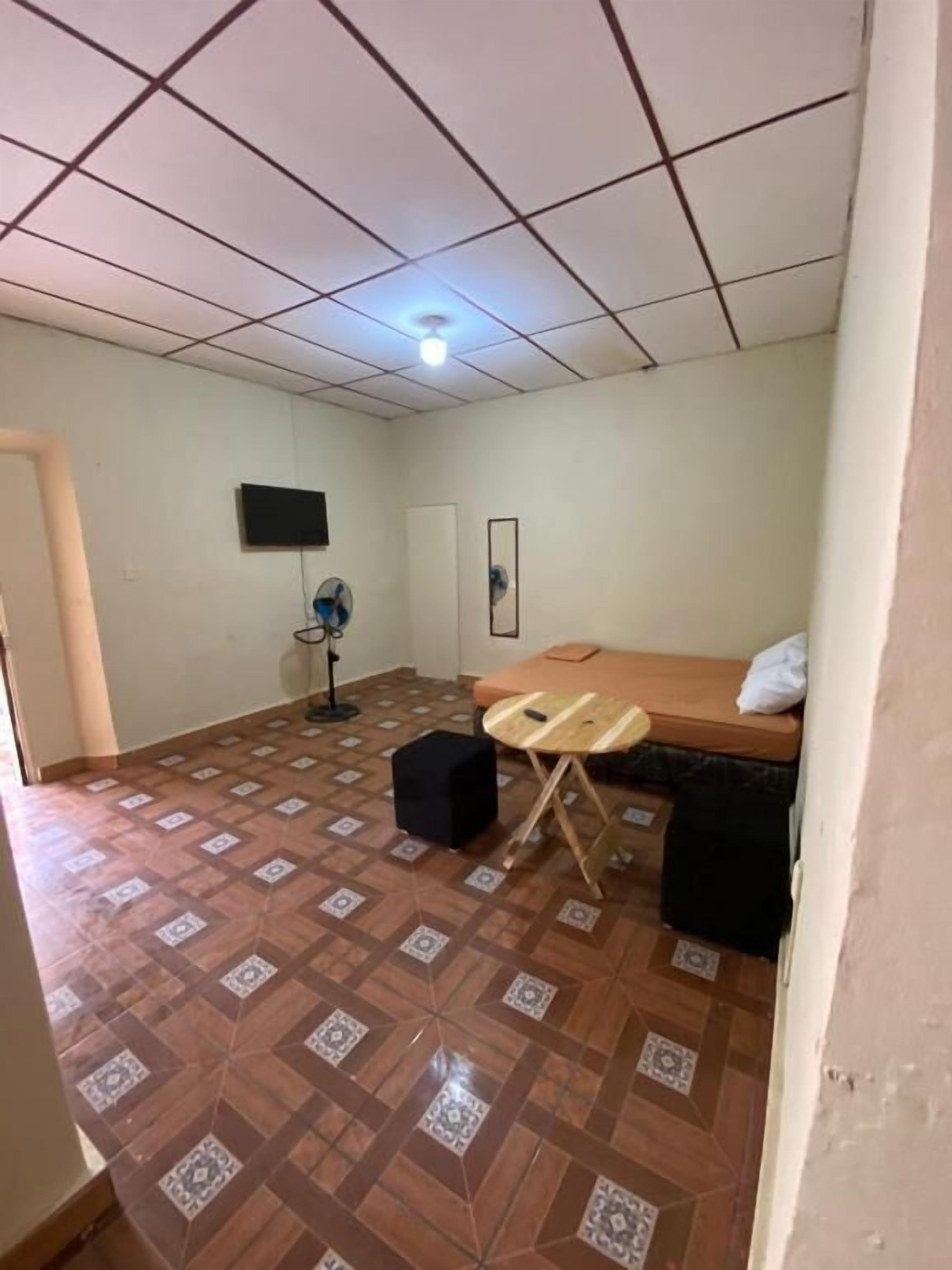 Une pièce avec un sol carrelé, un lit, un bureau, une chaise, un ventilateur et un téléviseur fixé au mur.