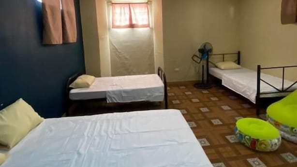 A room with two single beds, a window with curtains, a fan, and a small table.