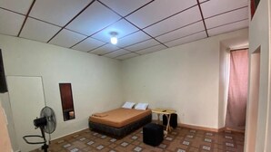 Una habitación con cama, ventilador de pie, una mesita y una ventana con cortinas.