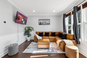 Ein Wohnzimmer mit Ecksofa, Flachbildfernseher, Stadtbildgemälde und einer Topfpflanze.
