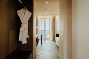 A hotel hallway with a robe hanging on a rack, a wooden door, a bench, and a bed with a view of the city.