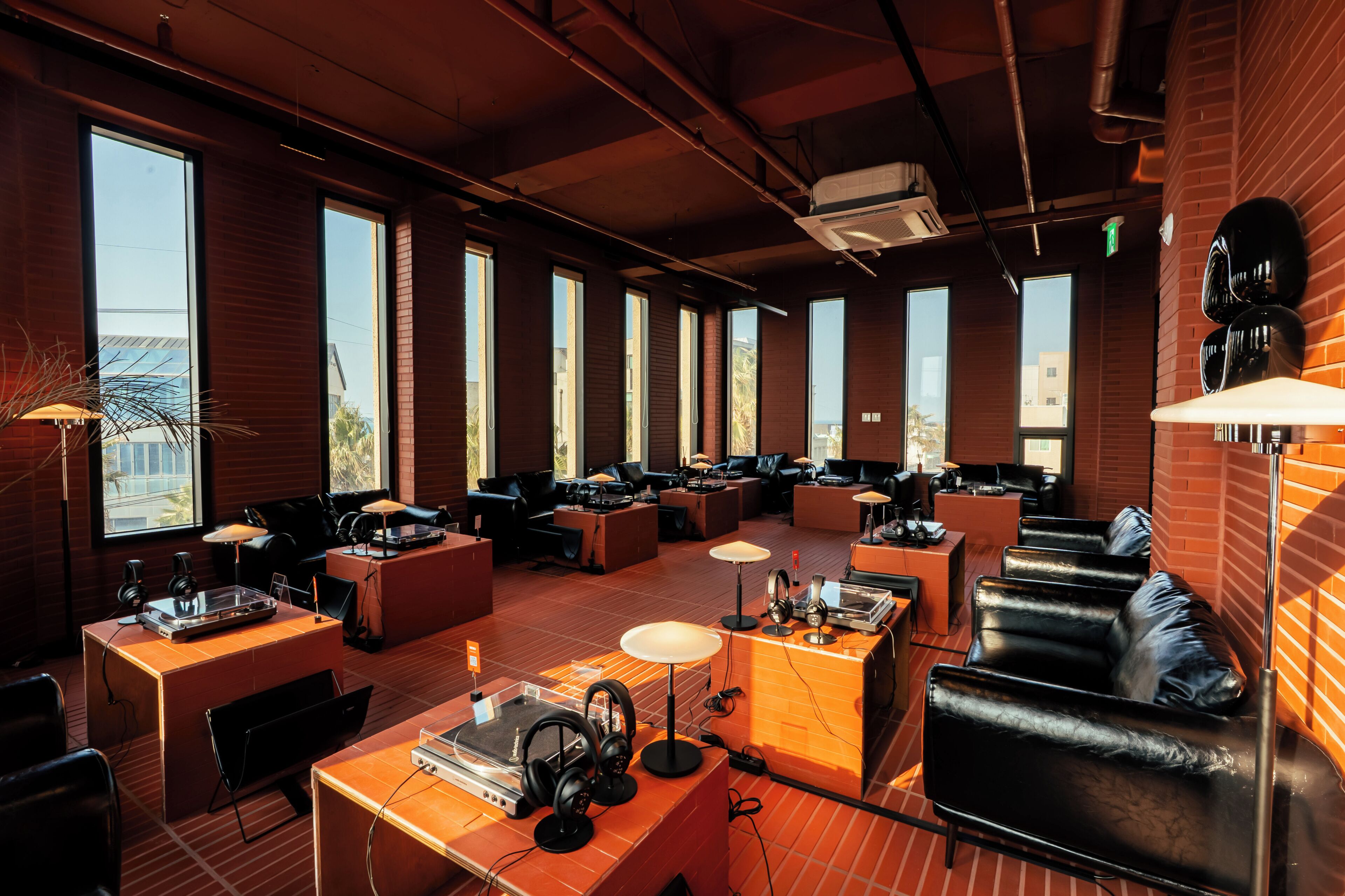 Amplia sala de estar con diversas opciones de asientos, incluyendo sofás y sillones, dispuestos alrededor de mesas con equipo de audio. Ventanas grandes ofrecen iluminación natural y vistas al exterior.