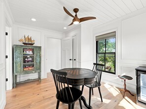 A dining area with a wooden table and chairs, a ceiling fan, a bookshelf, and a window with a view of greenery. - Historic Swansboro Retreat- 3-bedrooms- Water Views & Spa Bath (Swansboro)