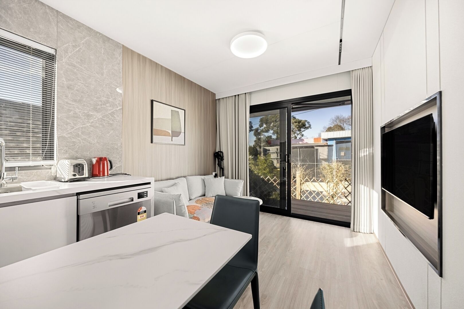 A modern kitchen with a dining area, a TV mounted on the wall, and a sliding glass door leading to a balcony.