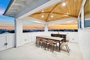 An outdoor patio with a wooden ceiling, a dining table with chairs, and a view of the cityscape at sunset. - Drift Away: Private Beach, Heated Pool & Spa, Golf Cart, Game Room, Rooftop View (Miramar Beach)