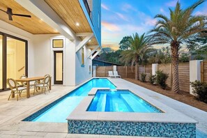 A swimming pool with a tiled border, a dining area with chairs, and a wooden deck. - Drift Away: Private Beach, Heated Pool & Spa, Golf Cart, Game Room, Rooftop View (Miramar Beach)