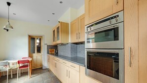 A kitchen with wooden cabinets, a stainless steel oven, and a dining area with a table and chairs. - 3BR Victorian Home for Up to 5 Guests (West Northamptonshire)
