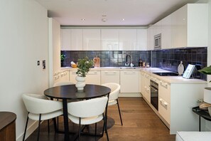 A modern kitchen with white cabinets, a black backsplash, and a round dining table with chairs. - 2BR Duplex Flat near Buckingham Palace (London)