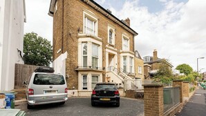 Une rue résidentielle avec deux voitures garées, une maison en briques dotée d’une baie vitrée et une clôture blanche.
