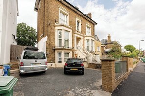 A residential street with two parked cars, a brick house with a bay window, and a white fence. - Bright 1BR Flat Opposite Peckham Rye Park (London)