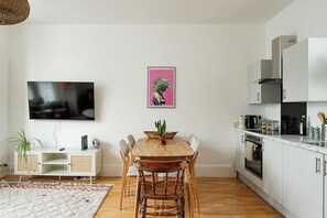 A modern kitchen with white cabinets, a wooden dining table, and a flat-screen TV mounted on the wall. - Bright 1BR Flat Opposite Peckham Rye Park (London)