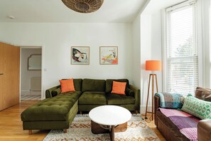 Ein modernes Wohnzimmer mit einer Eckcouch, einem runden Couchtisch und einer Stehlampe. An der Wand hängen zwei gerahmte Kunstwerke, und ein Fenster mit Jalousien.
