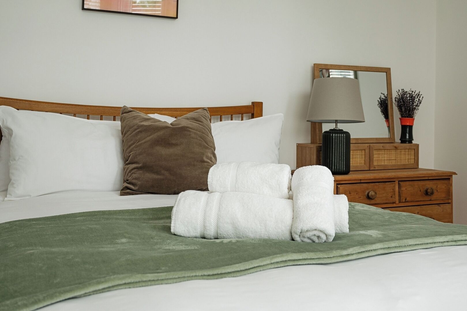 A neatly made bed with white and brown pillows, a green throw blanket, and rolled white towels on a wooden headboard.