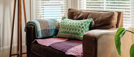 A brown leather sofa with a colorful blanket and patterned pillows, positioned in front of a window with white blinds.