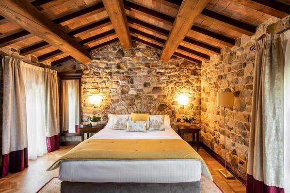 Un dormitorio con pared de piedra, vigas de madera, una cama con almohadas y dos lámparas fijadas a la pared.